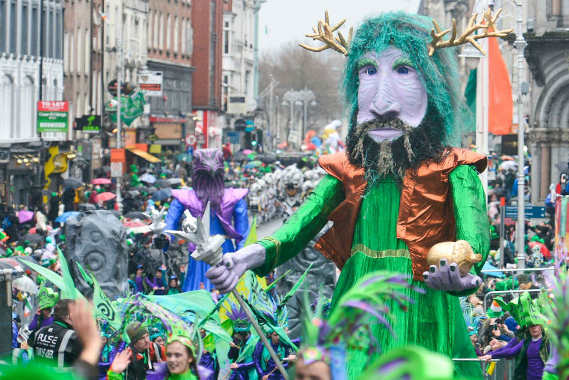 A shot of the colourful parade in Dublin with a huge paper mache model of a man with purple skin, green hair and golden horns holding a golden acorn.