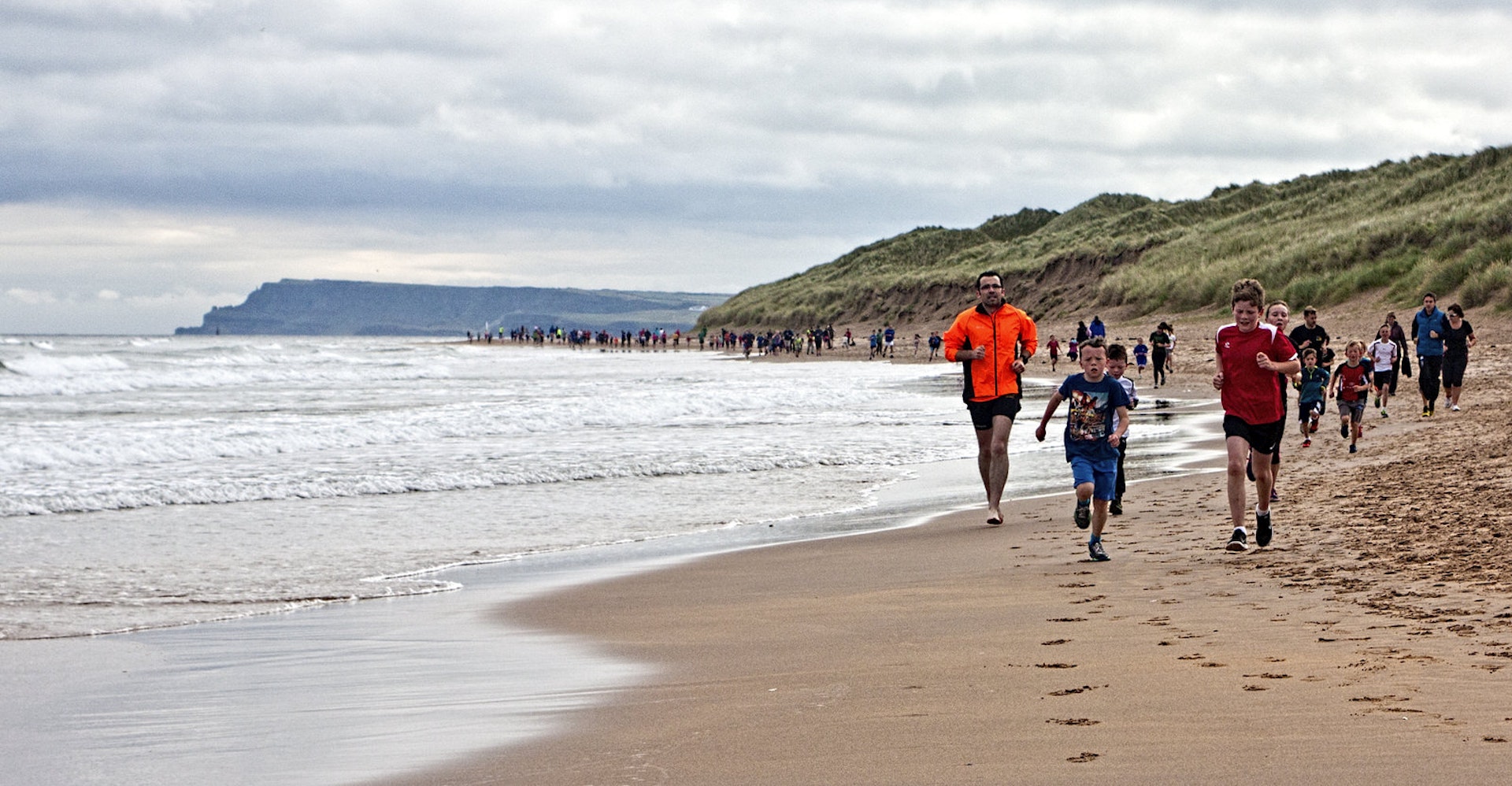 Runners, approaching the camera, stretch out along the beach in Portrush; at the front are some children, with a middle-aged man in bright orange running barefoot