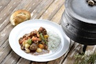 A bread roll sits on a wooden-plank table next to a white plate topped with white rice and a traditional ste of potatoes, carrots, greens and beef; sat next to both is a traditional three-legged cast iron pot (aka potije)