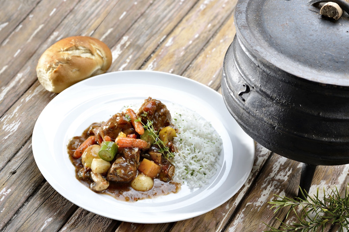 A bread roll sits on a wooden-plank table next to a white plate topped with white rice and a traditional ste of potatoes, carrots, greens and beef; sat next to both is a traditional three-legged cast iron pot (aka potije)