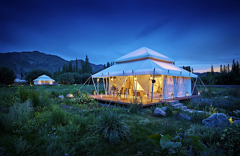A square white tent with two-tired pyramidal roof glows with soft light in the dusk with the mountains of Ladakh in the background; the tent sits amongst lush vegetation, another similar tent is in the distance