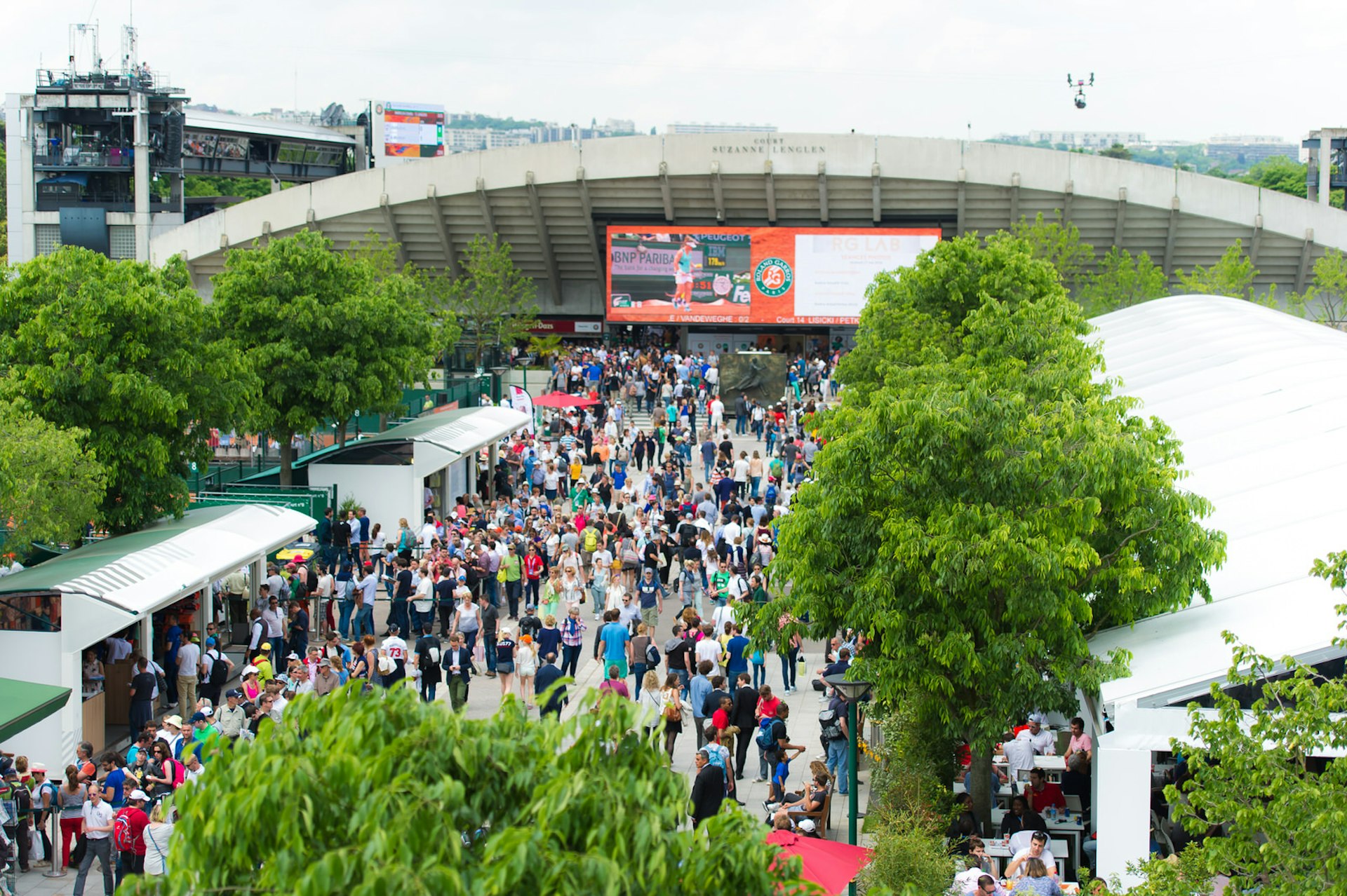 Crowds in colourful attire walk to and fro along a wide boulevard leading up to Court Suzanne Lenglen; on either side of the pedestrian path are temporary stalls beneath large green trees selling drinks, food and tennis paraphernalia