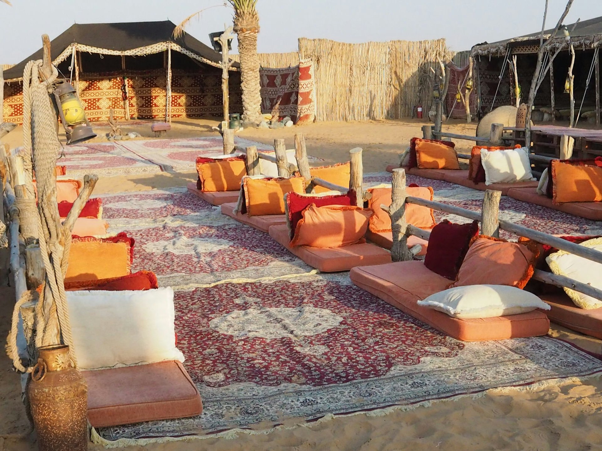 A camp is set up in the desert in Dubai, with patterned red-and-white carpet and low, cushioned seats set up on the sand. 