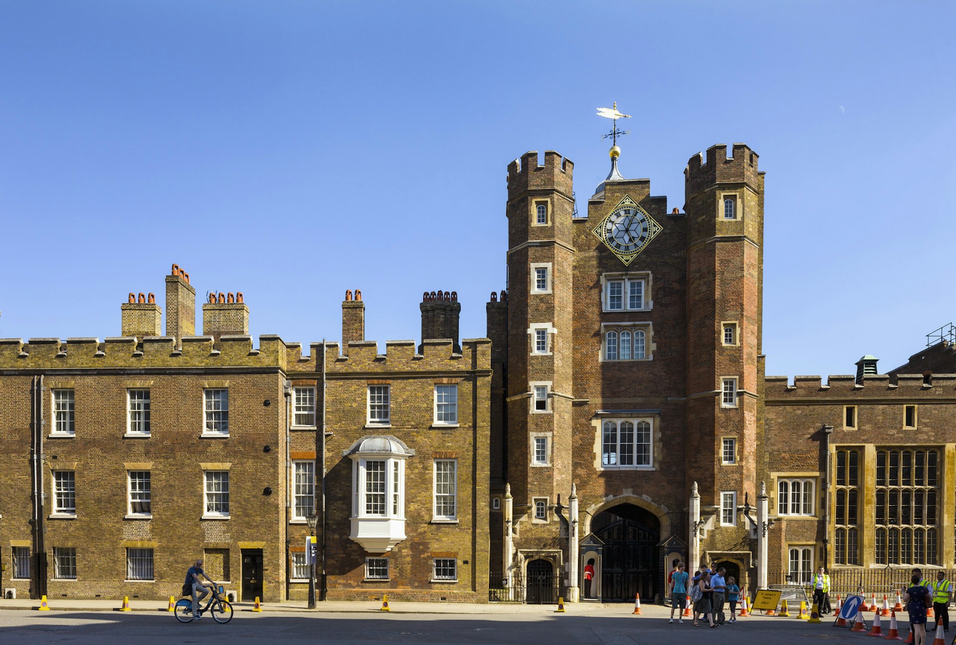 The brownish redbrick facade and narrow castellated entrance towers of St James's Palace are dotted with neat little windows with white window bars; high above the entrance is a large gilded clock © Photolibrary / Getty Images Plus