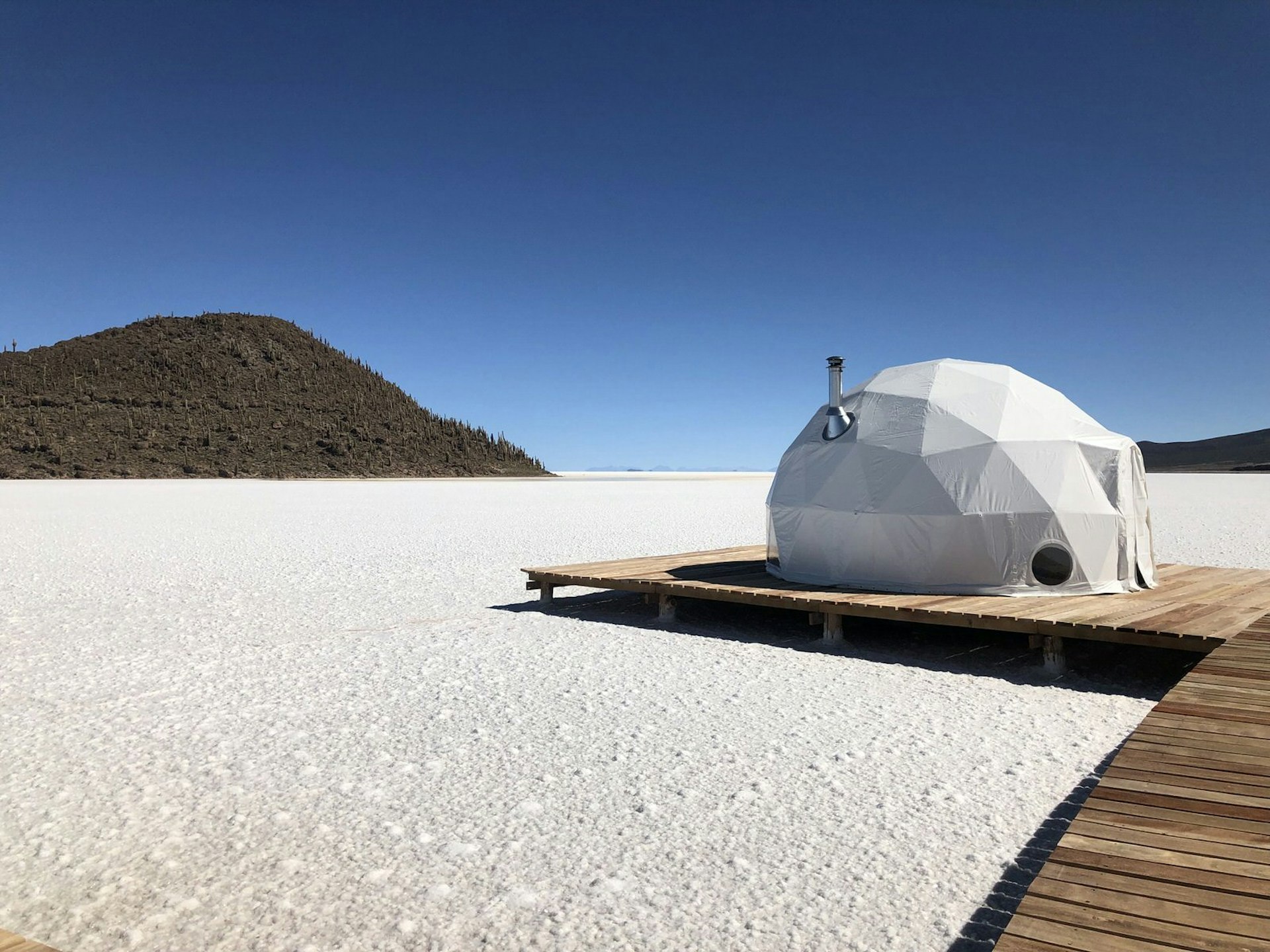 A white geodesic domed tent with a silver chimney sits on a raised wooden platform, which seemingly hovers over white salt flats; in the semi-distance is a forested hill rising from the flats, much like an island