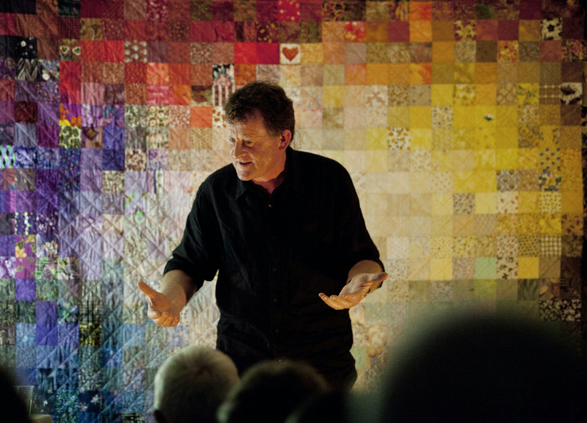 A man in black top and pants tells stories with his palms open to the audience, a multi-coloured patchwork quilt as a backdrop behind him