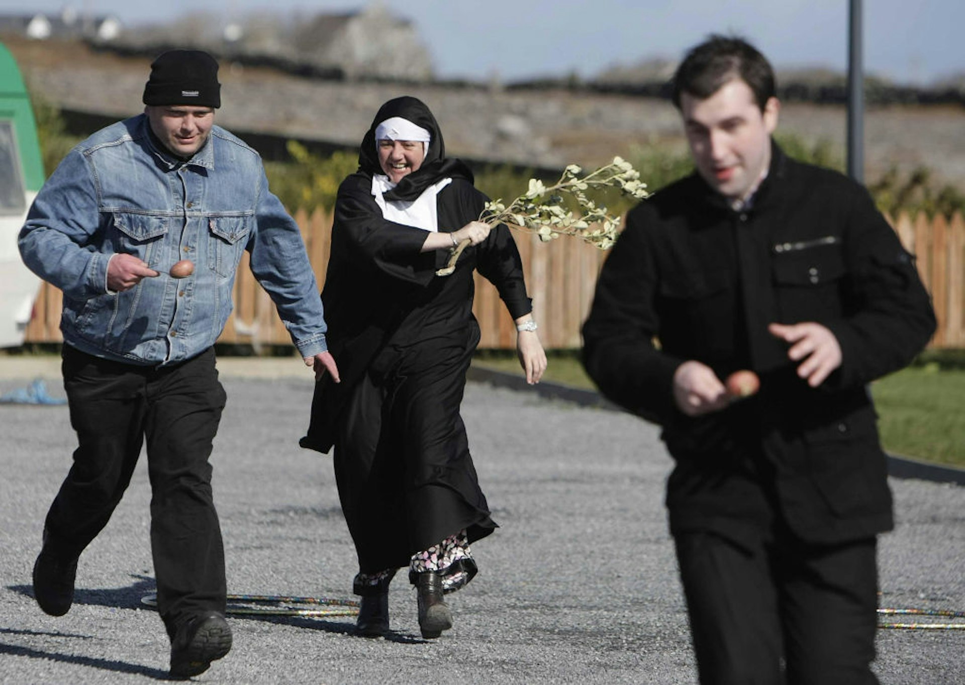 A woman dressed as a nun waves a stick behind two men in an egg and spoon race.