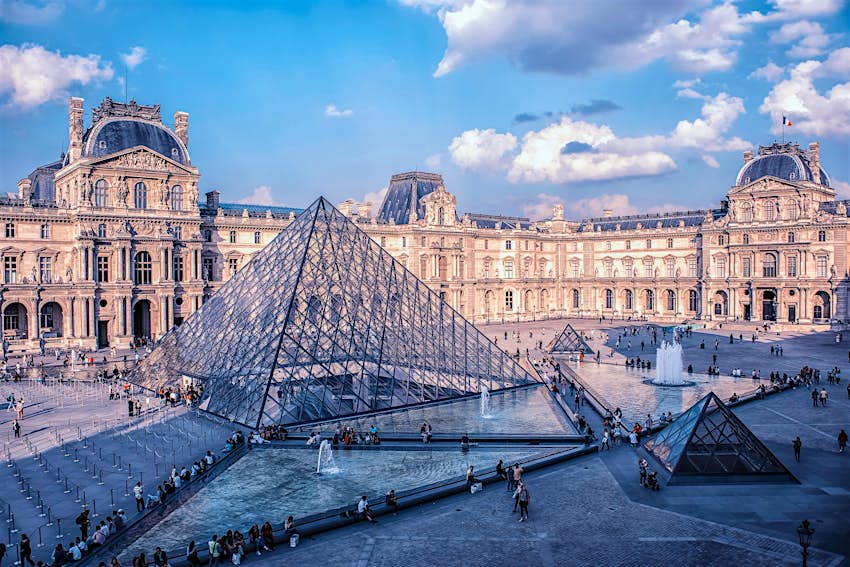 The large glass pyramid which serves as the entrance to the Louvre Museum surrounded by fountains and figures;  the backdrop is the gloriously detailed facade of the historic palace
