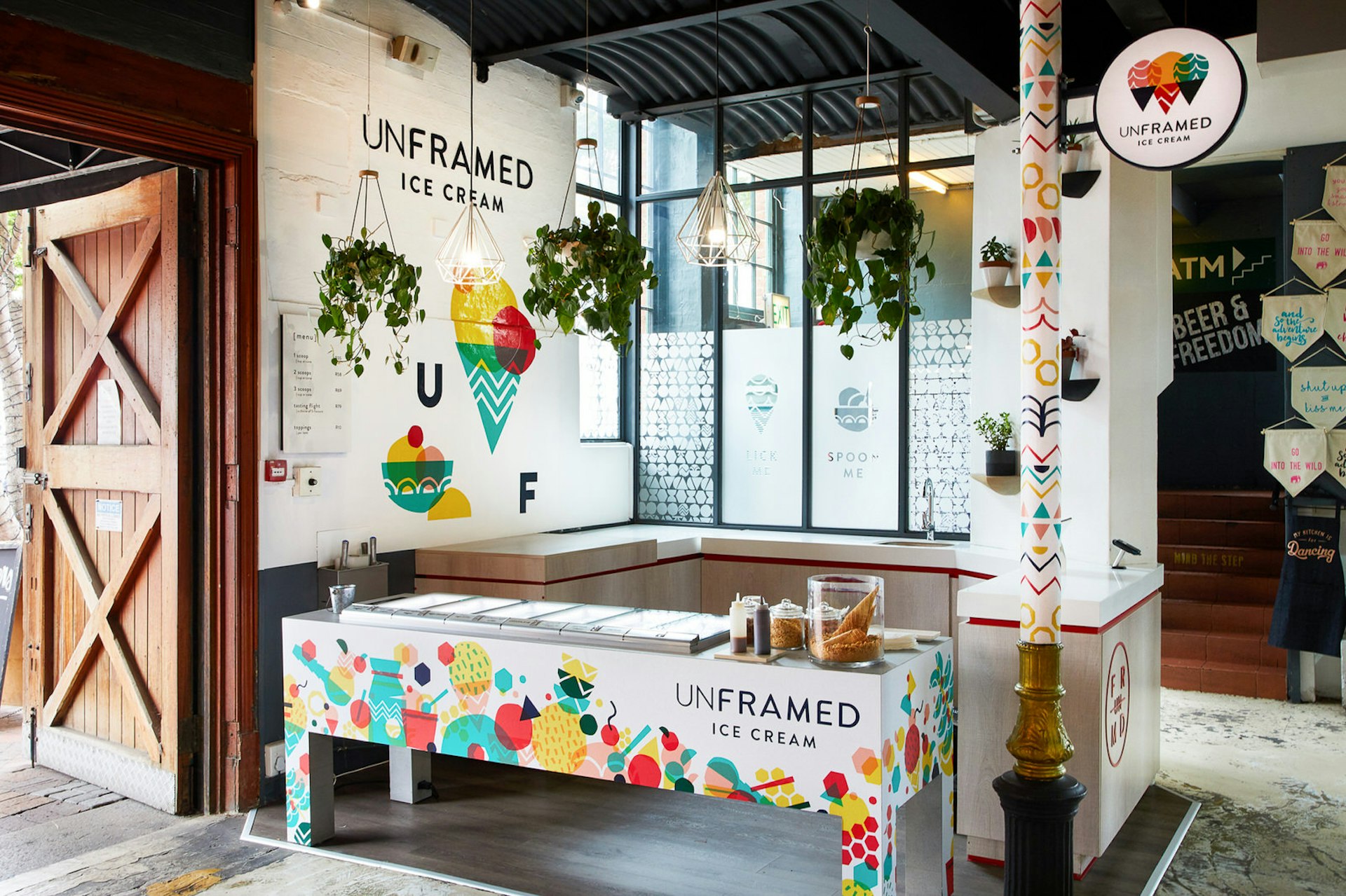 A large barn-like wooden door hangs open and lets light fill the bright interior of Unframed; white walls are covered in vibrant murals of abstract cones and ice cream scoops, with a dark grey ceiling high above