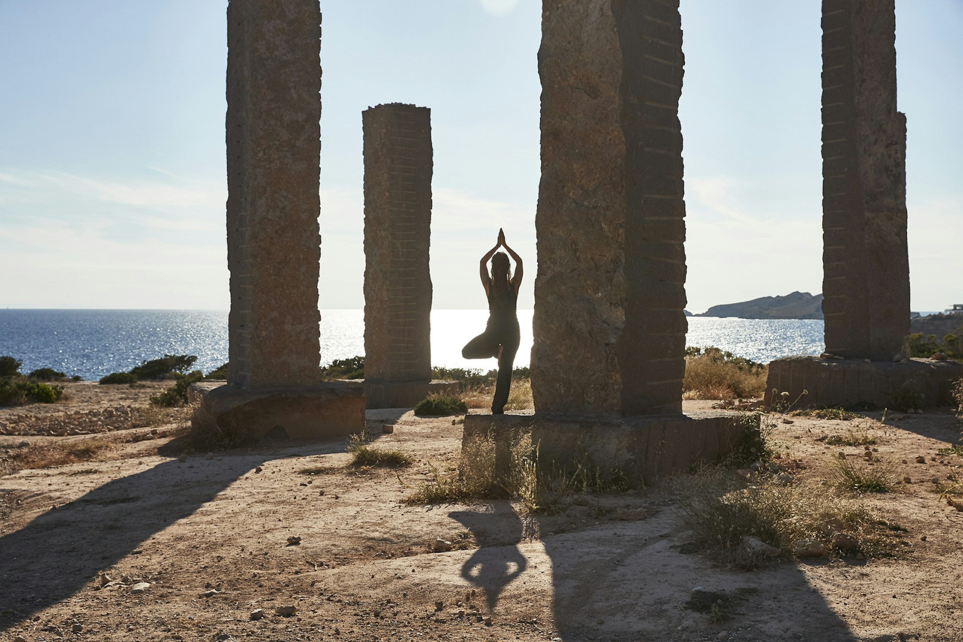 Ibiza spa breaks - a person in tree pose next to four tall vertical rock pillars overlooking the sea