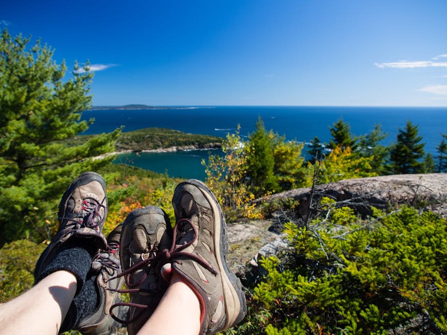 Hiking boots are stretched out over a coastline view as people are resting at the top of a mountain in Acadia National Park