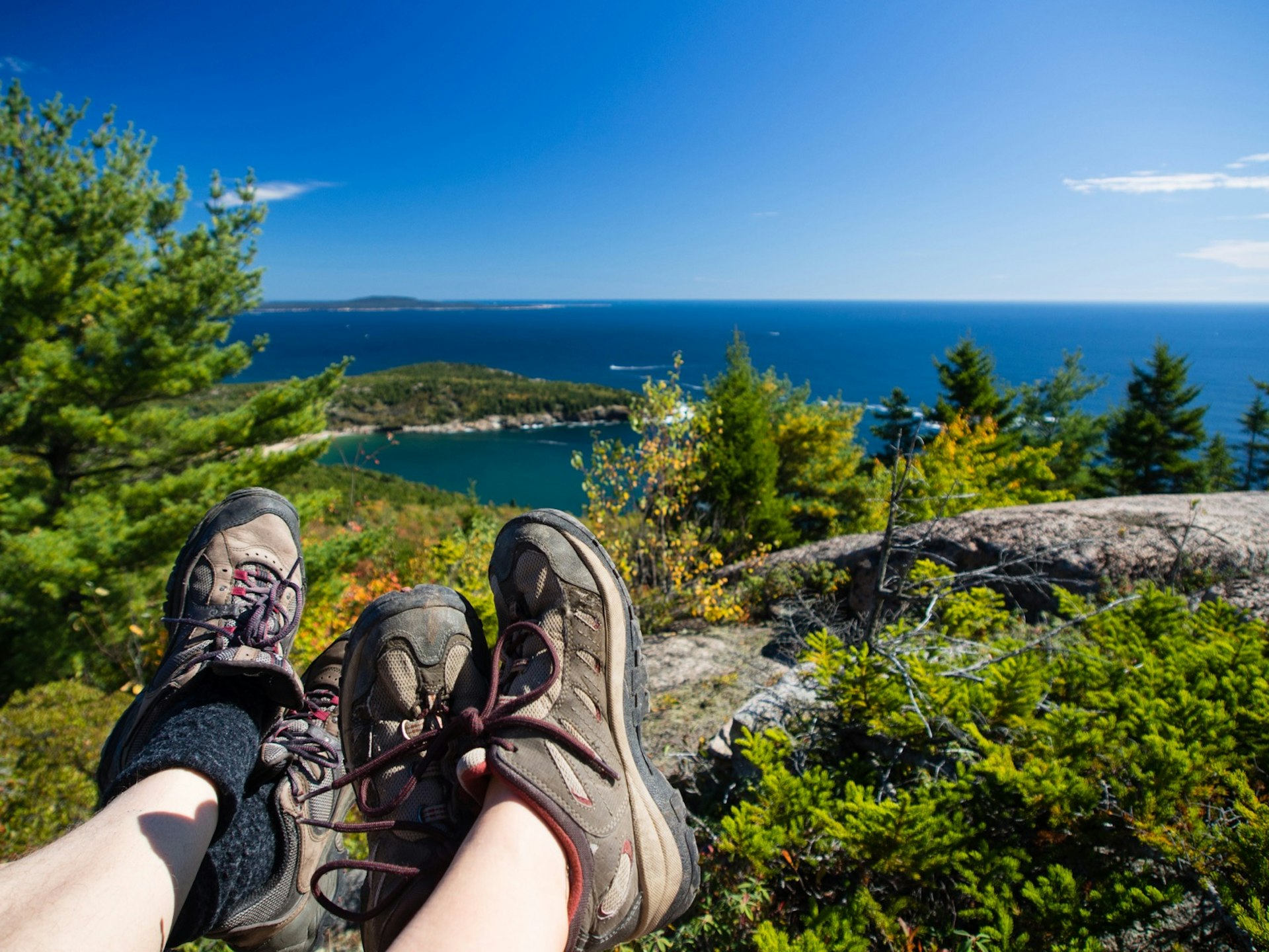 Hiking boots are stretched out over a coastline view as people are resting at the top of a mountain in Acadia National Park