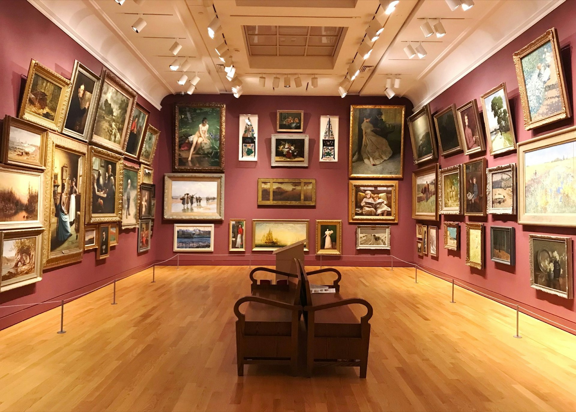 Rich maroon walls and blonde wood floors mark a museum gallery with dozens of framed artworks on every wall