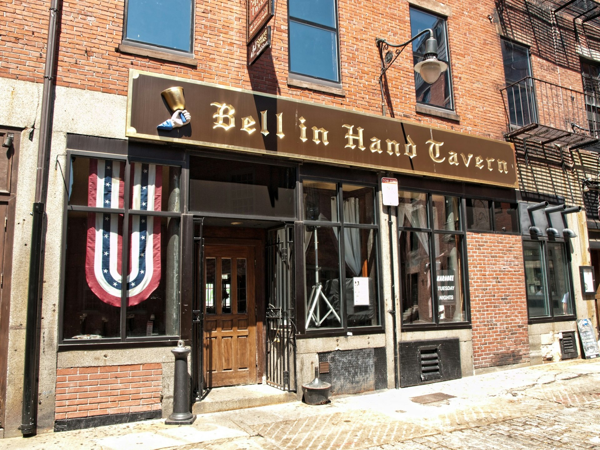 A long brown sign with an image of a hand and a bell mark this brick pub as the historic Bell in Hand Tavern
