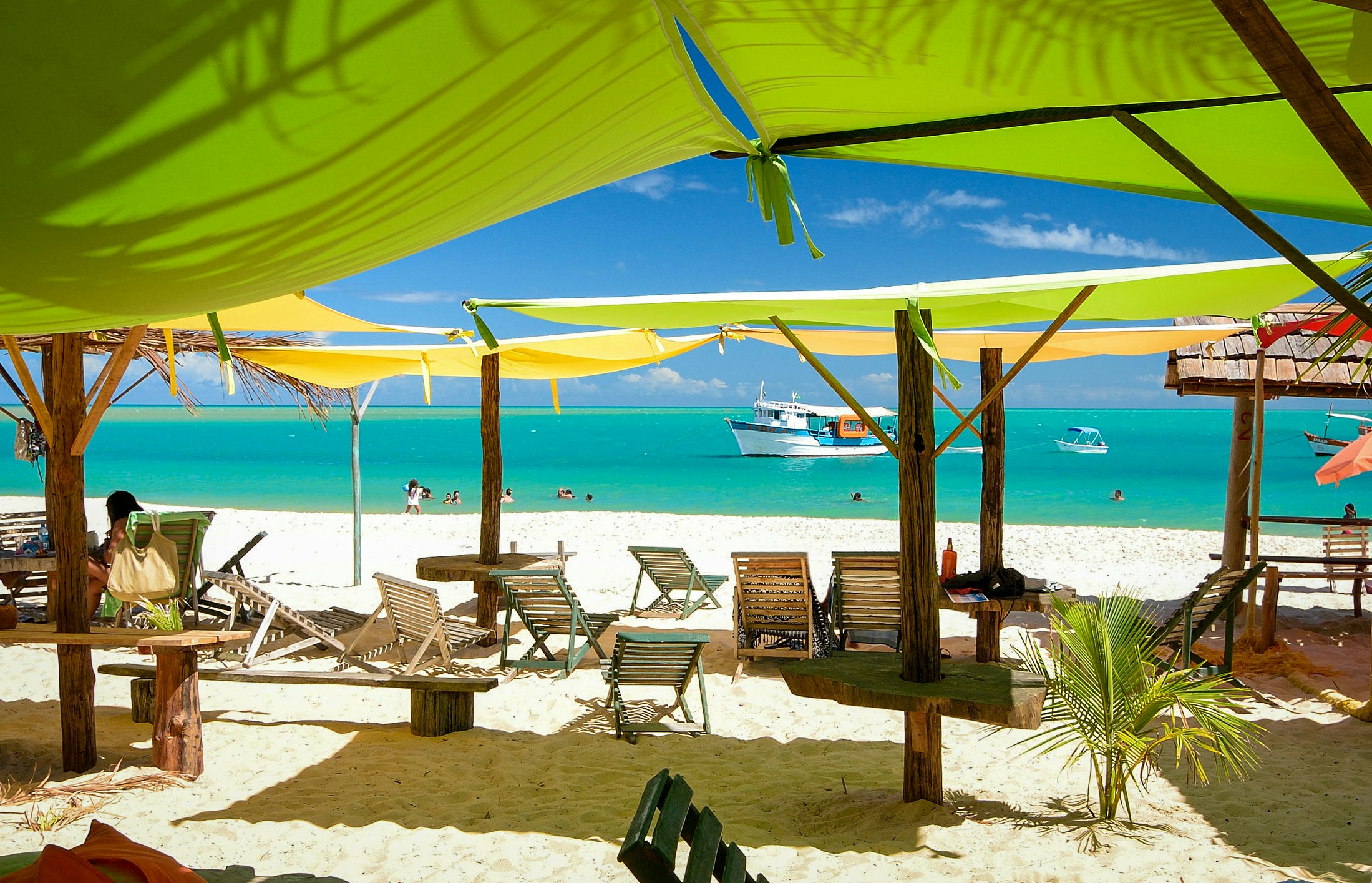 Wooden beach loungers sit on white sand underneath green and yellow awnings; bright blue water and a boat are visible in the background. Bahia, Brazil.