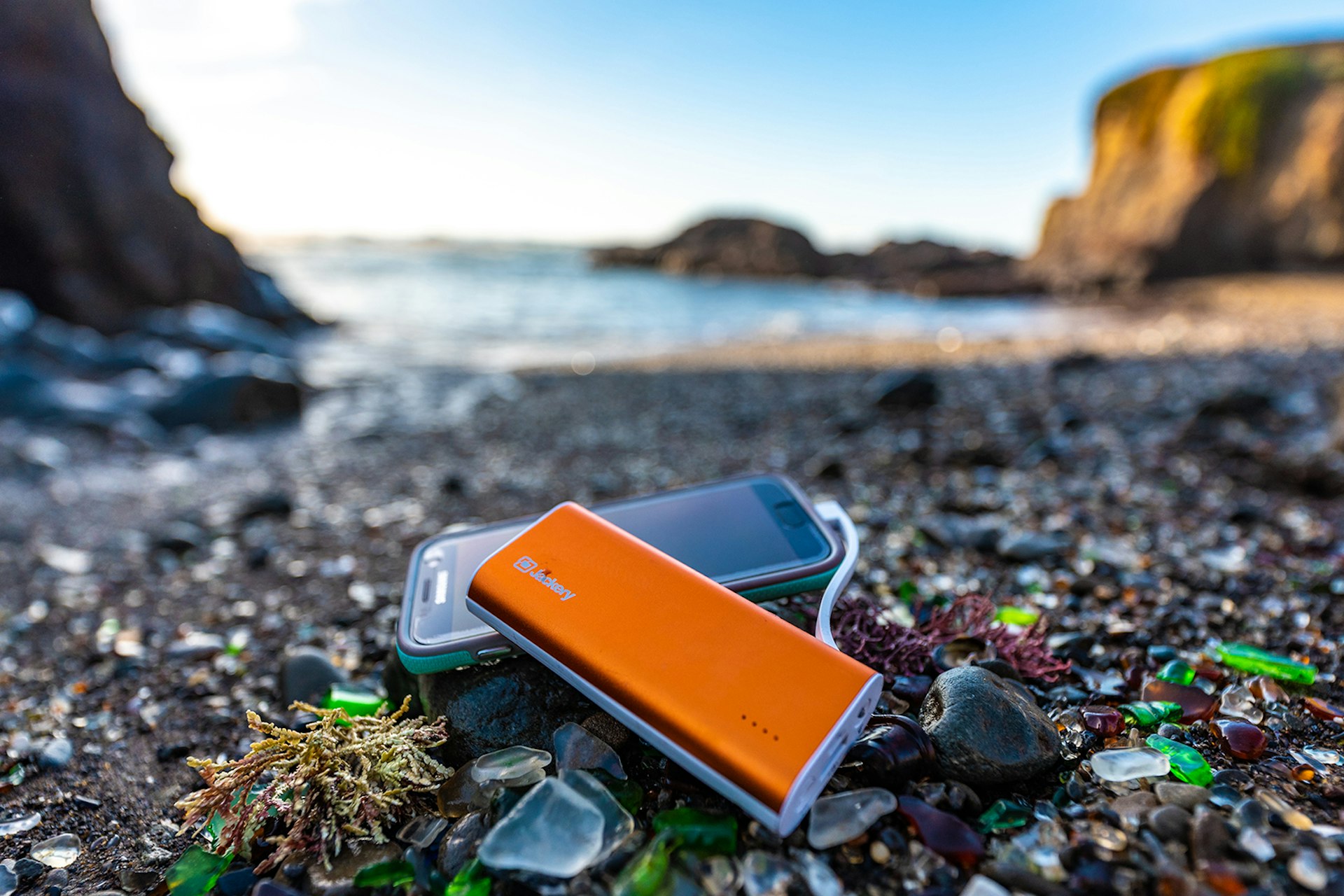 Product shot of the Jackery’s Bolt 6000 and a smartphone. The focus is on the orange charger and the beach which is littered with cut glass. The blurry sea and cliffs are visible in the background; Father's Day gifts