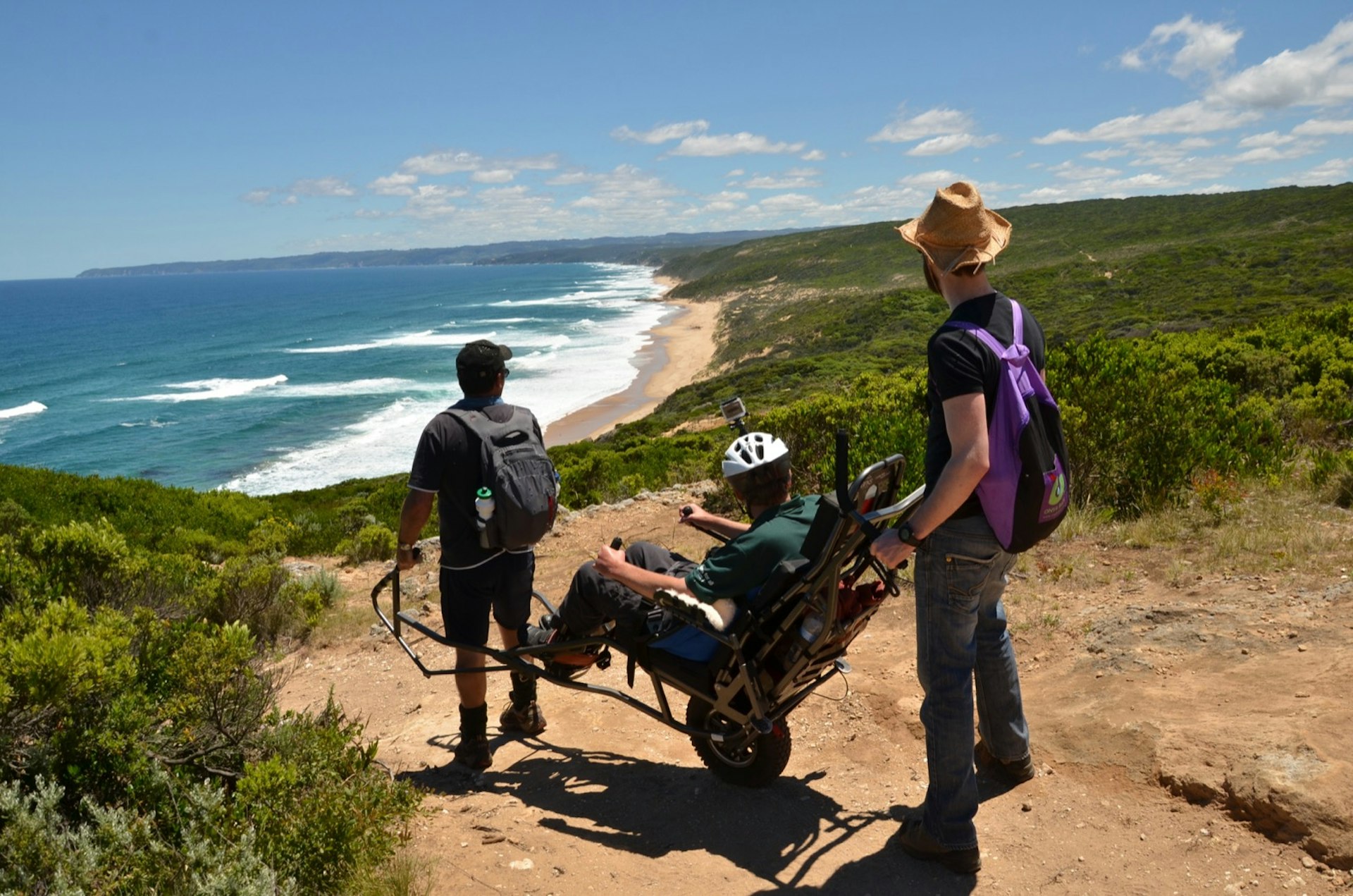 Three people are on a bluff over looking the ocean, one is in a two-wheel wheel chair, one holds the front and the other stands at the back of the wheelchair working to make Australia Accessible; accessible Australia