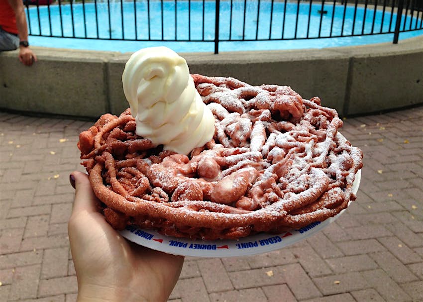 A hand holds out a funnel cake, cooked to a deep brown, with a dollop of soft-serve ice cream on top of it.