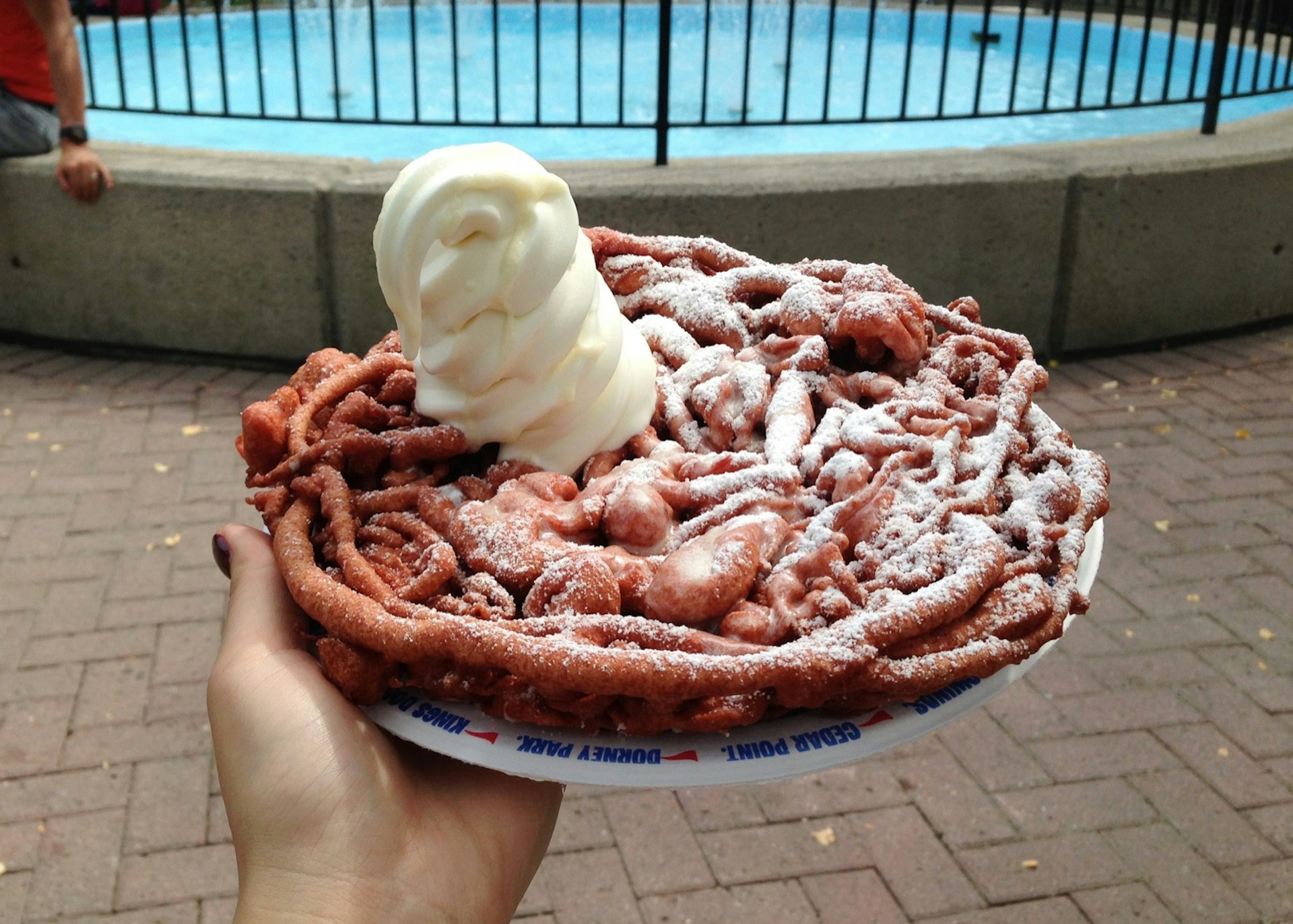 A hand holds out a funnel cake, cooked to a deep brown, with a dollop of soft-serve ice cream on top of it.