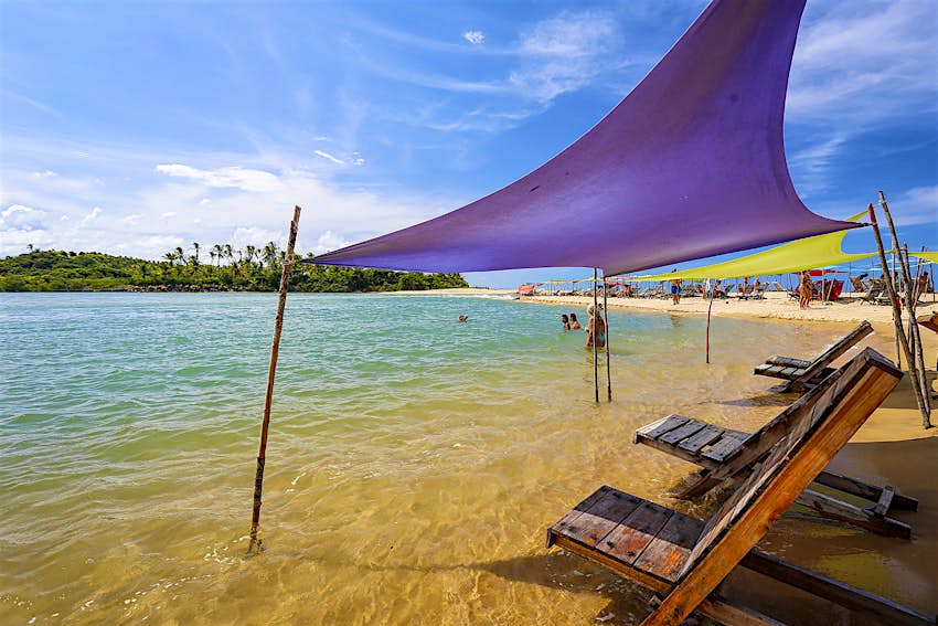 Features - Caraiva , Bahia , Brazil Wooden beach loungers sit under purple and green awnings on a sandbar in Caraíva. Bahia, Brazil.