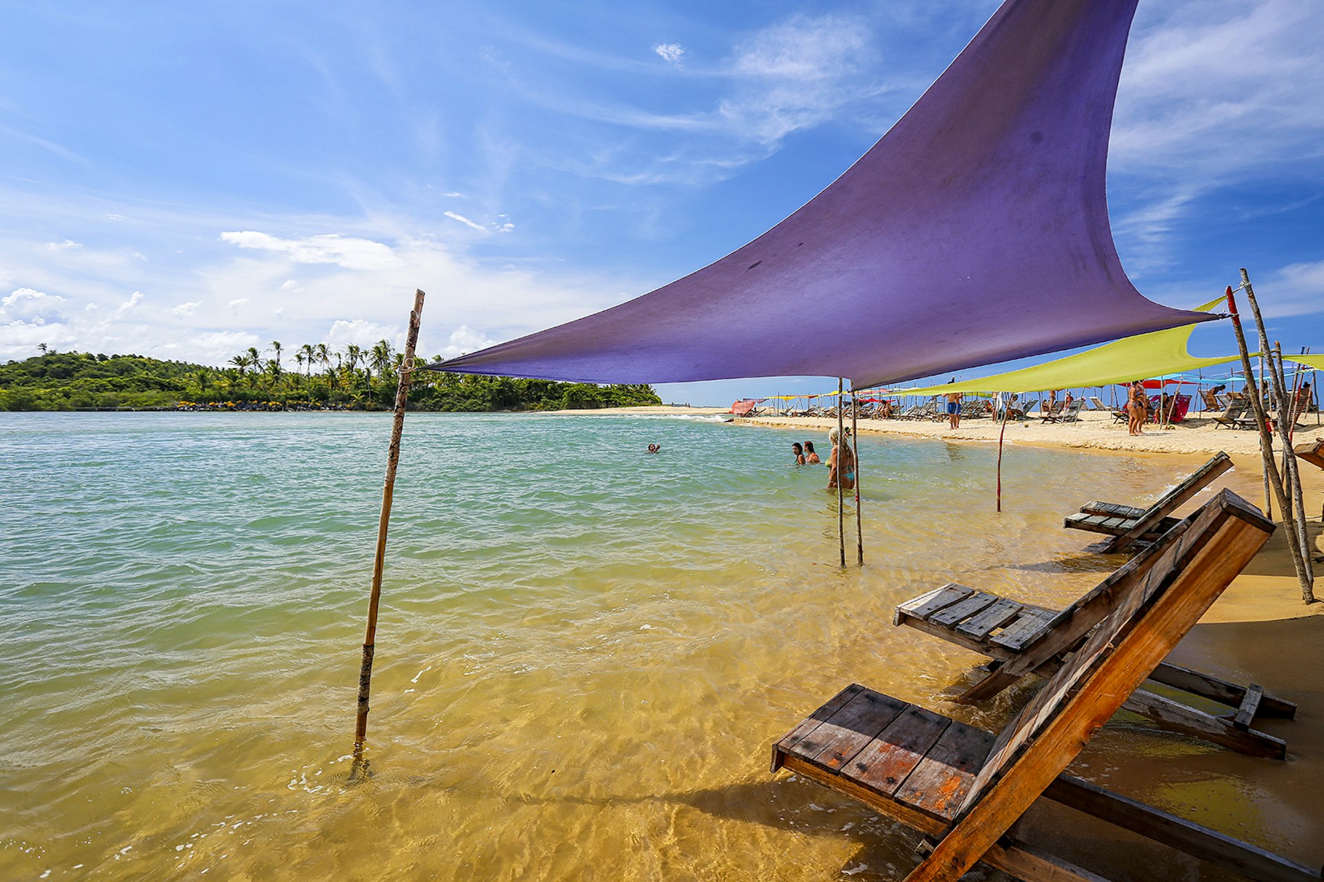 Wooden beach loungers sit under purple and green awnings on a sandbar in Caraíva. Bahia, Brazil.
