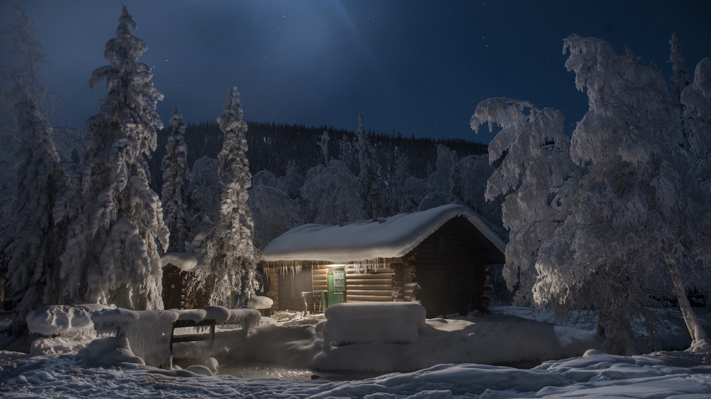 A snow-covered cabin and trees surround a water hole at night at Chena hot springs near Fairbanks Alaska.