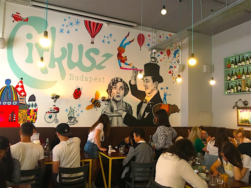 Diners cram into a high-ceilinged cafe with light bulbs hanging from long wires; the back wall is covered in a vibrant mural of balloons, flying trapeze artists, planes and ladybirds