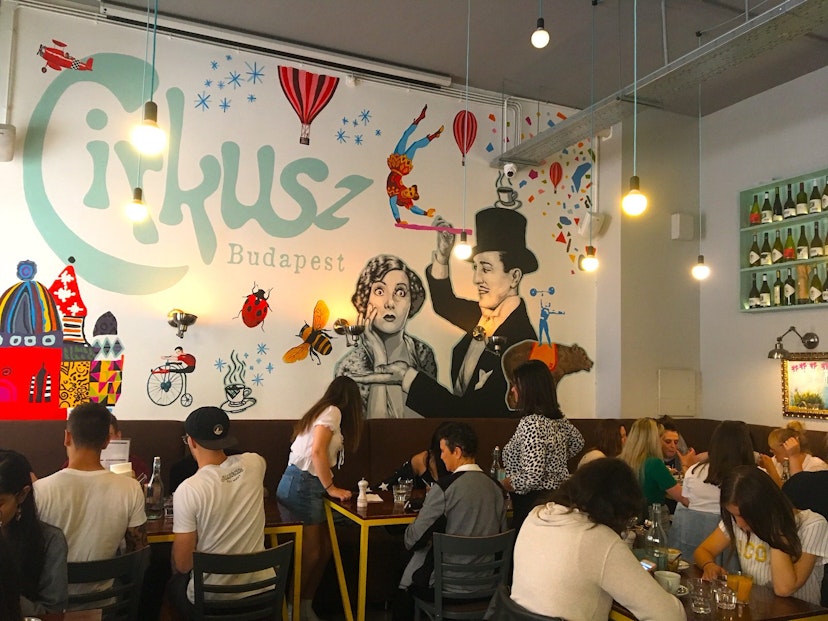 Dinners cram into a high-ceilinged cafe with light bulbs hanging from long wires; the back wall is covered in a vibrant mural of balloons, flying trapeez artists, planes and lady birds