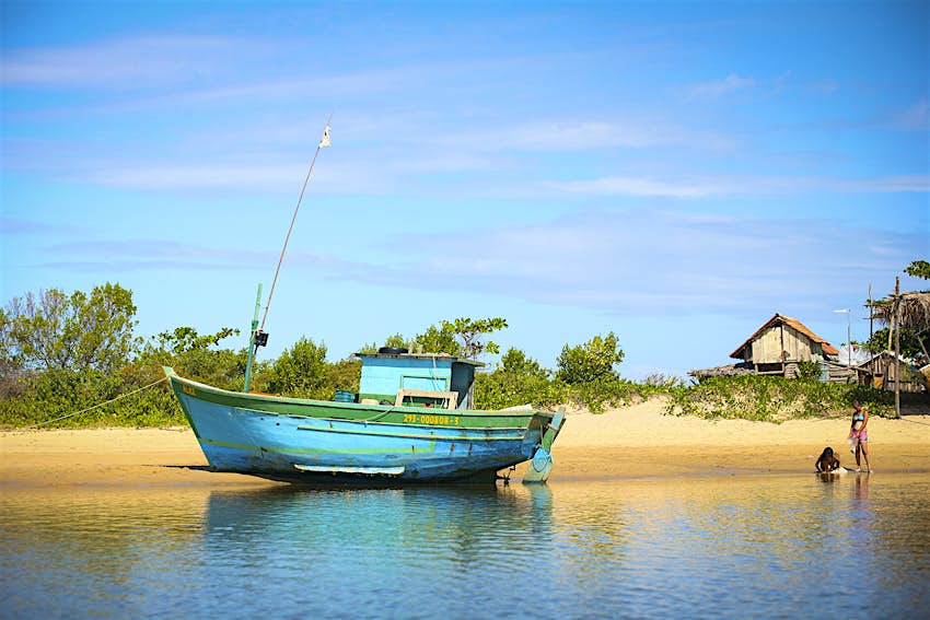 A blue wooden fishing boat sits beached on a golden beach, with a fishing shack in the background.