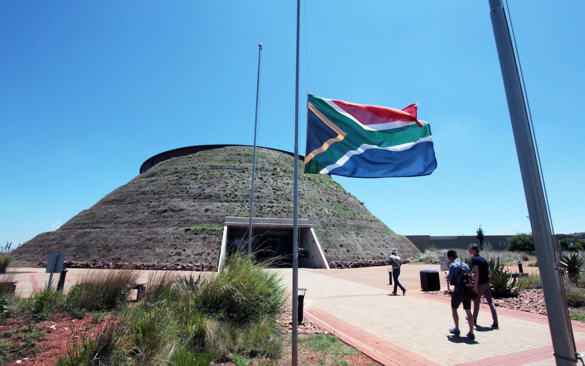 A couple of visitors walk beneath a flowing South African flag and towards a large grass-covered dome; above it all is a brilliant blue sky