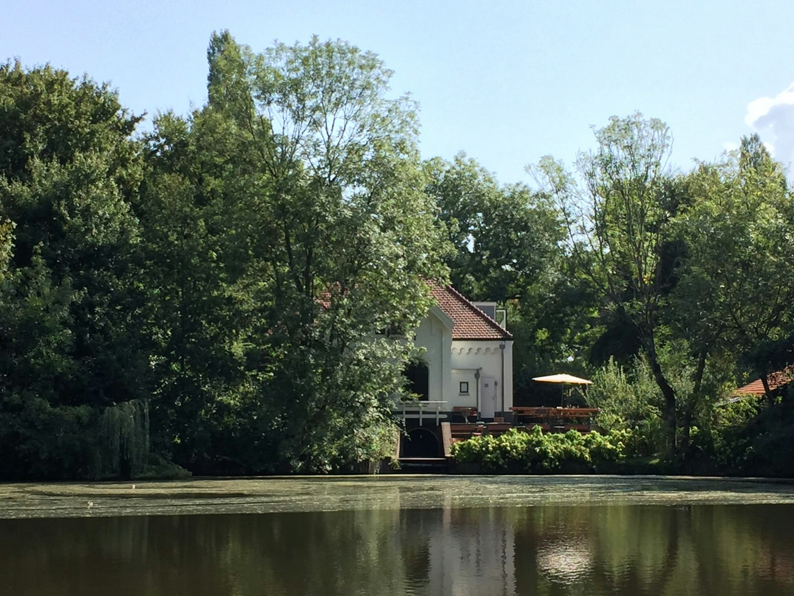 Looking across the water to an old pump house partly obscured behind trees; the sky is blue and beautiful, the water dead calm