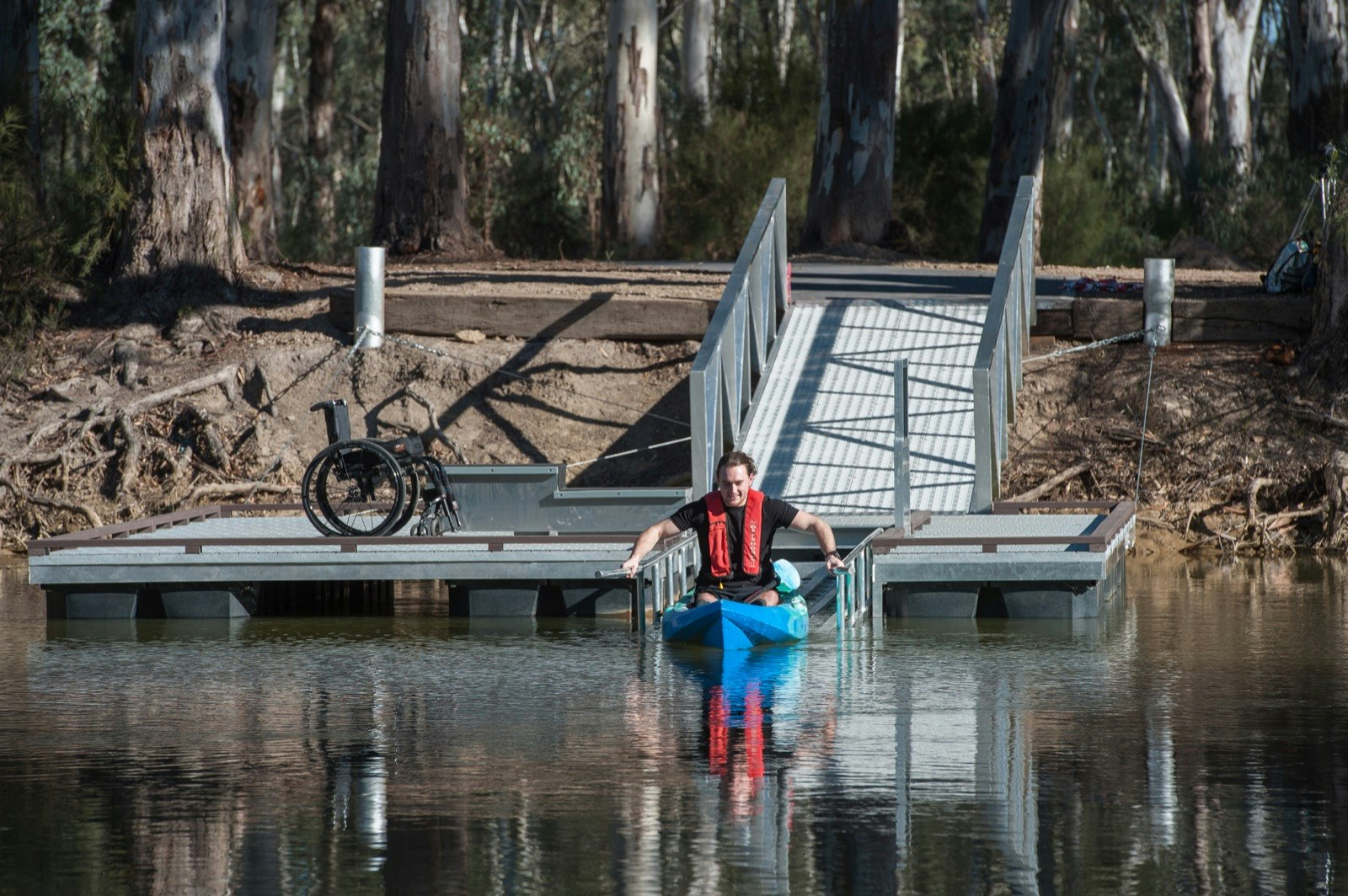 A man launches his kayak into the water while a wheelchair sits on a dock behind him; accessible Australia