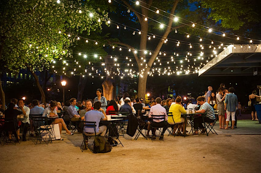 Numerous of tables and patrons sit beneath rows of golden fairy lights hanging from trees within Madidson Square Park