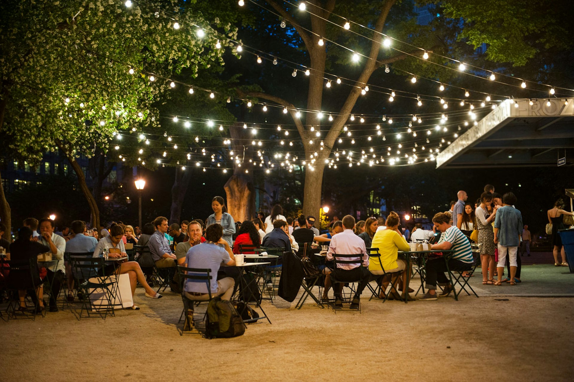 Numerous of tables and patrons sit beneath rows of golden fairy lights hanging from trees within Madidson Square Park