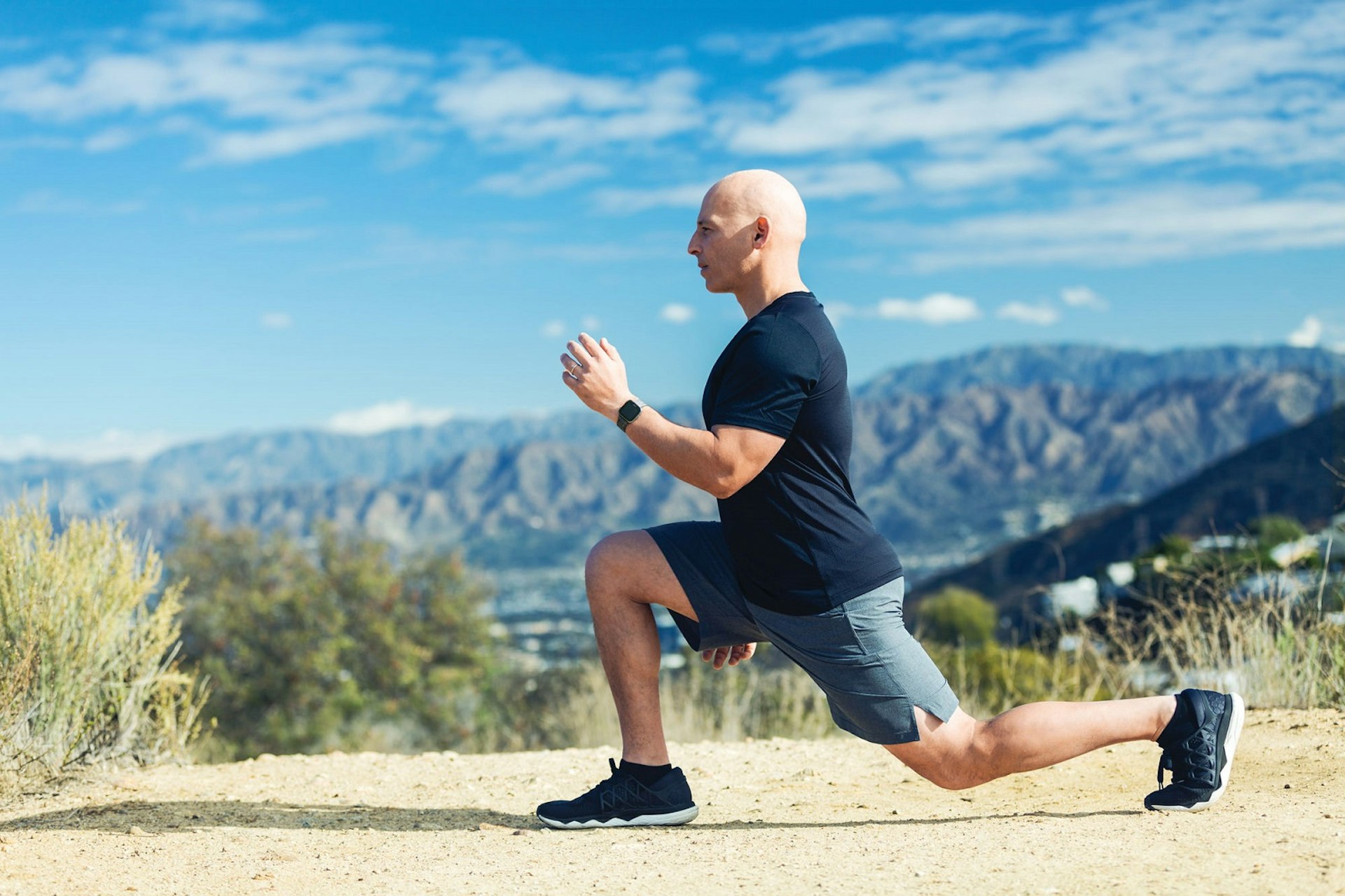 Lifestyle Photo of Harley Pasternak wearing a Fitbit watch. Harley is doing a reverse lunge on a hiking path with panoramic views of hills in the background