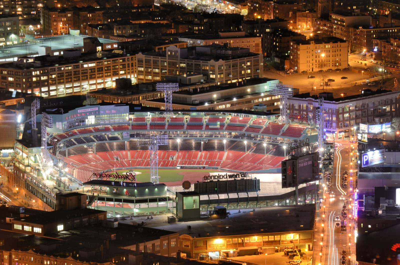 Aerial view of an illuminated Fenway Park at night. There are buildings surrounding the stadium and cars driving by.