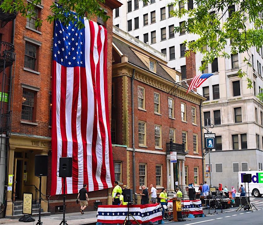 Features - Fraunces-Tavern-1-0a0163223f06 The historic brick building of the Fraunces Tavern is seen on a street corner in Manhattan, across the street from a skyscraper. A giant American Flag is hung across the facade of the building next door and a band sets up to perform outside the tavern