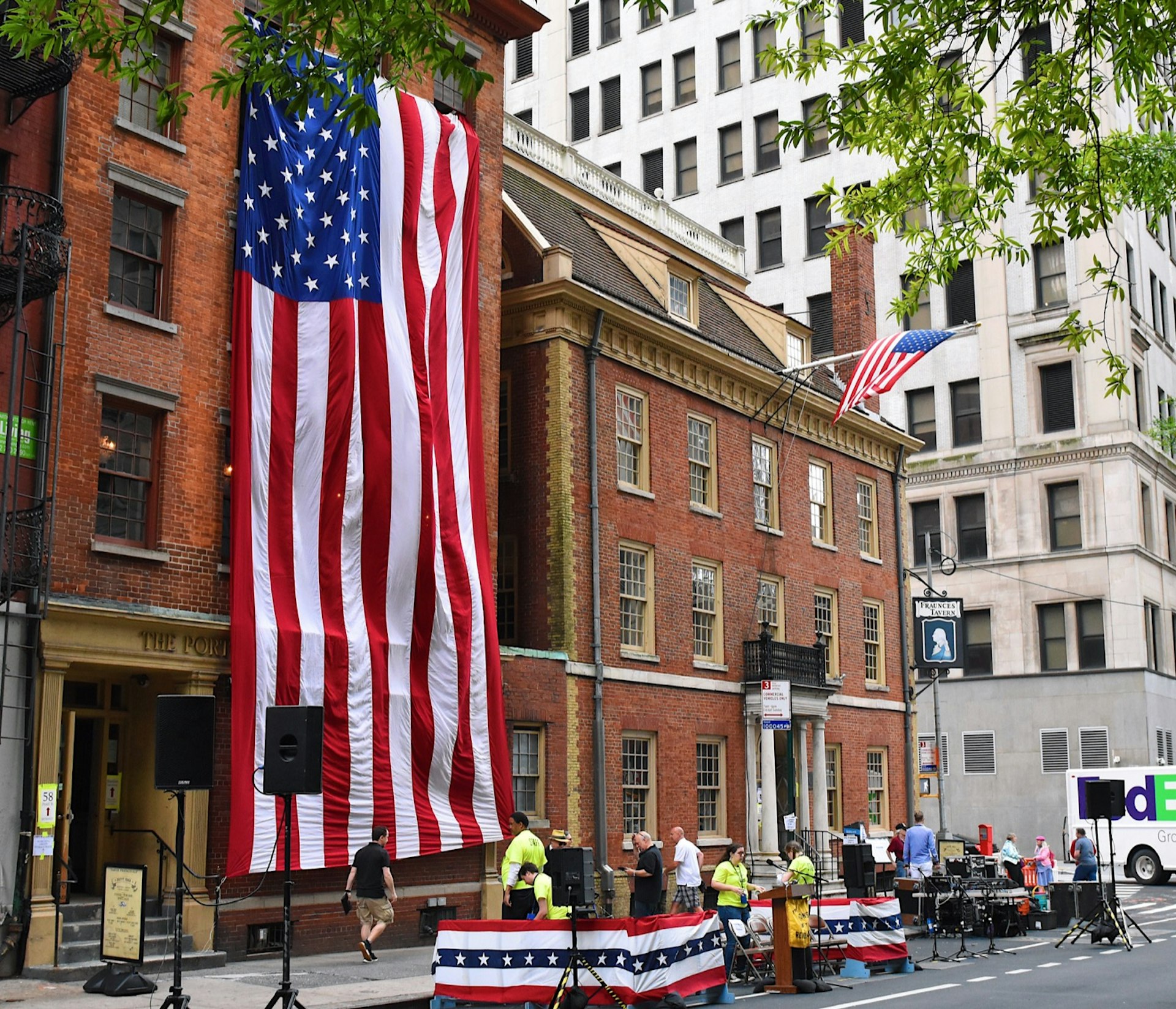 The historic brick building of the Fraunces Tavern is seen on a street corner in Manhattan, across the street from a skyscraper. A giant American Flag is hung across the facade of the building next door and a band sets up to perform outside the tavern