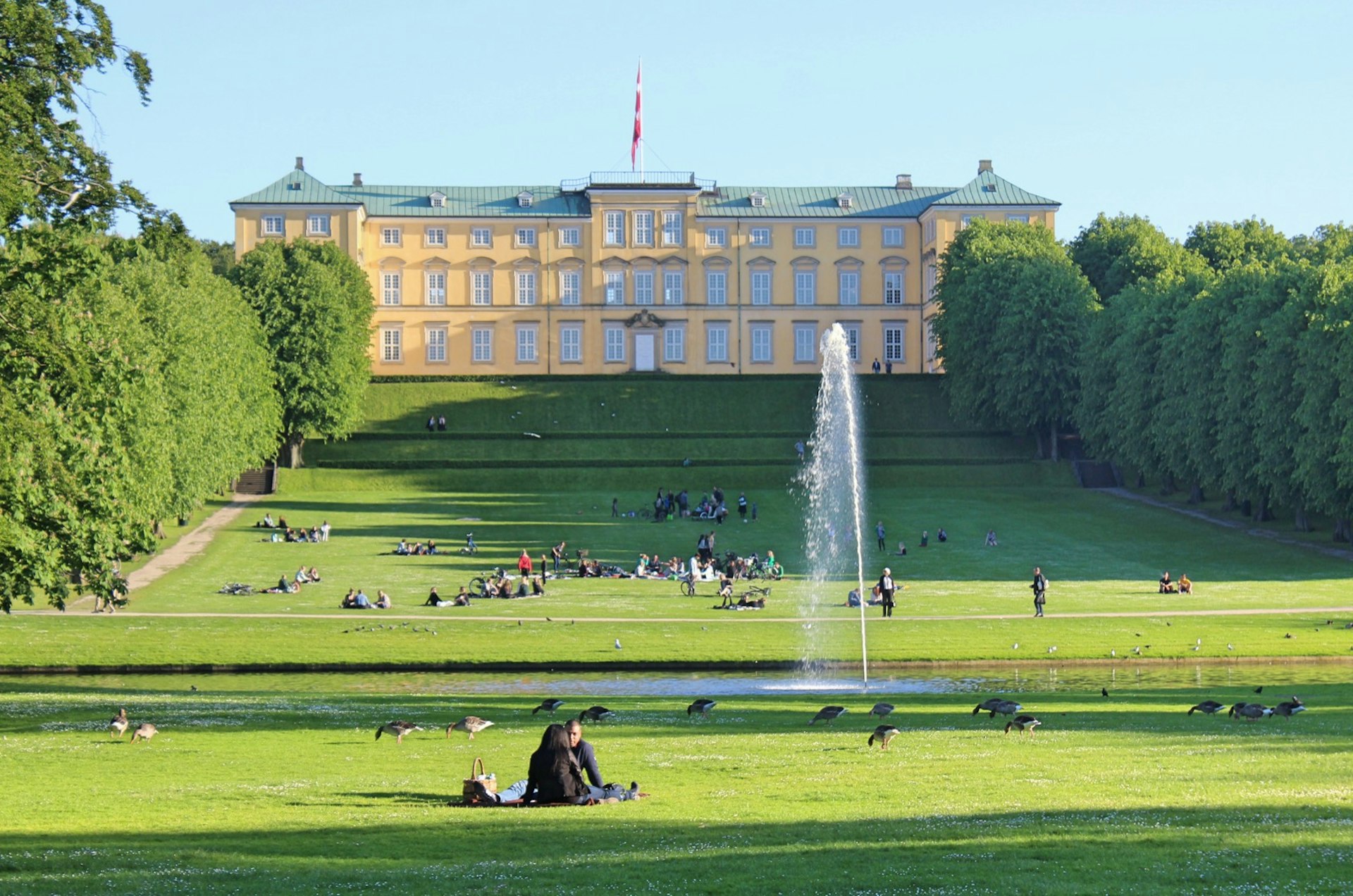 Groups of people sit scattered along the expansive grassy lawn and fountain in front of Frederiksberg Castle at Frederiksberg Have. The garden sits on 32-hectares of land. 
