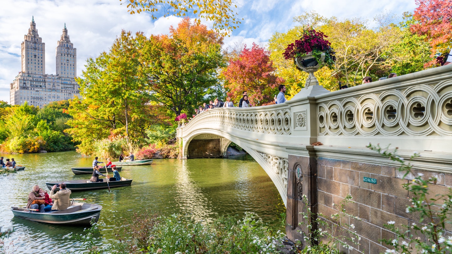 A stone bridge arches over a calm, narrow lake in Central Park; boats paddle to its left and all the trees are changing colours, with leaves green, yellow, orange and red