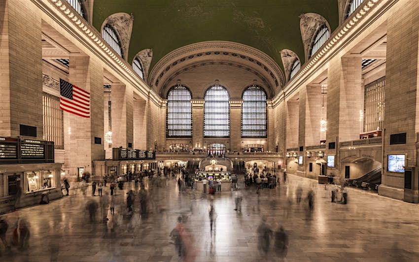 A view down the main hall of Grand Central Terminal; three large windows at the far end tower above the blurred masses of people moving to and fro; high above it all is a deep green ceiling painted with golden stars