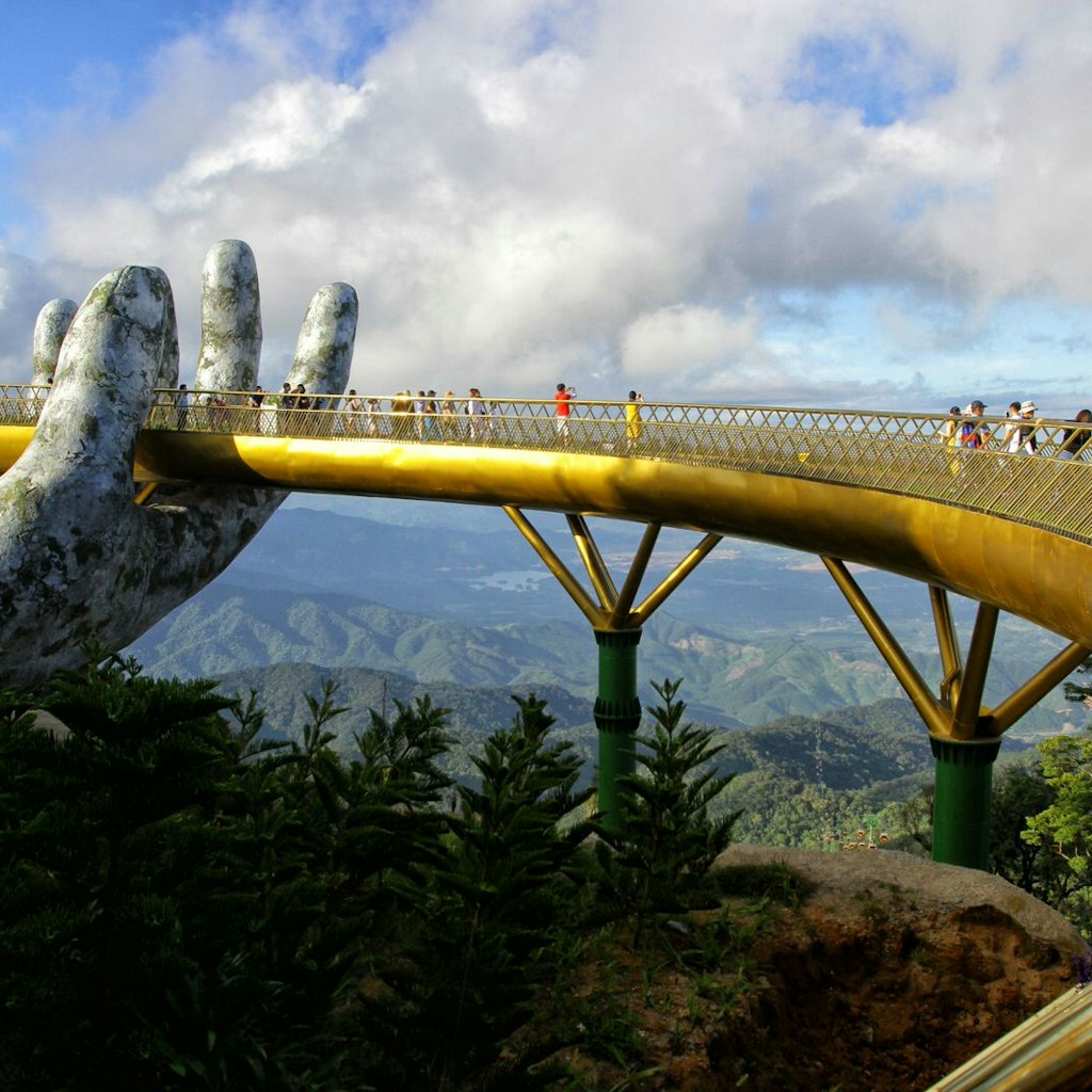 A single giant concrete hand can be seen holding the golden walkway of the bridge, as several people enjoy the view of jungle and hills that stretches for miles