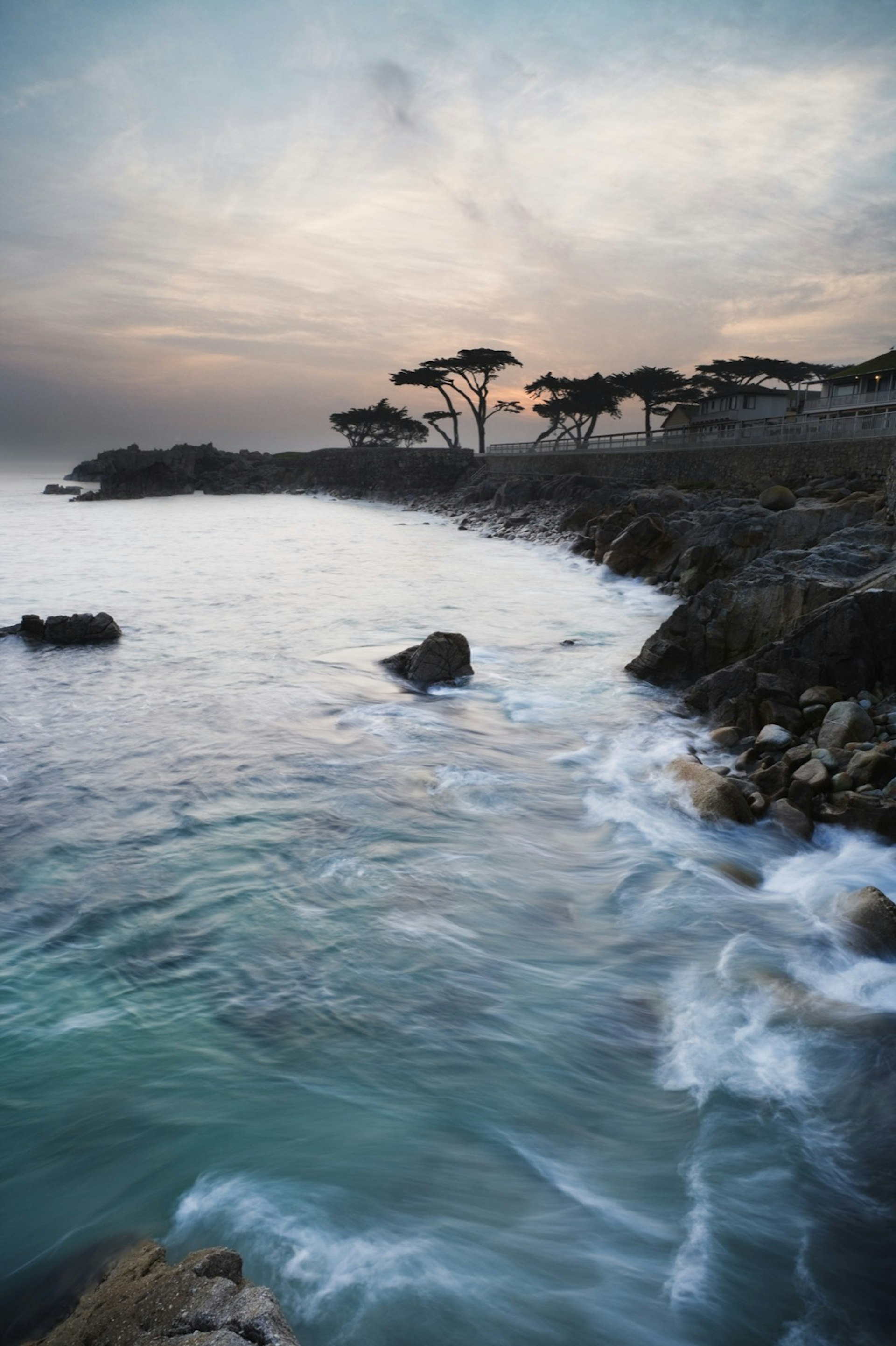 Waves crash on a rocky shore with the sun rising over cypress trees and a rocky peninsula