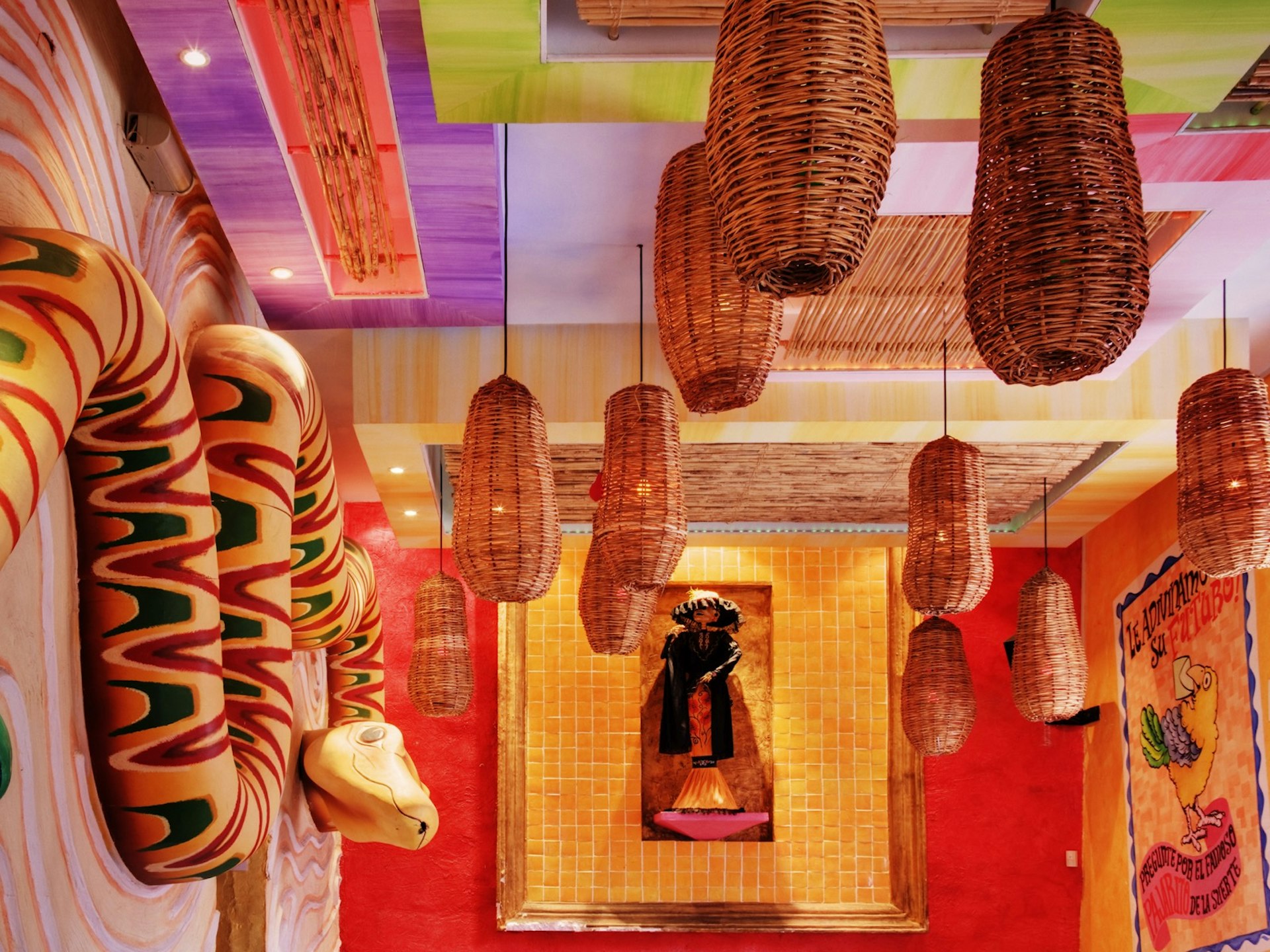 A painted wooden snake is affixed to the left wall while the back wall houses an image of a person in traditional dress the right wall has an image of a snake and woven baskets hang from the ceiling being used as lamps. It is painted purple, green, red and orange