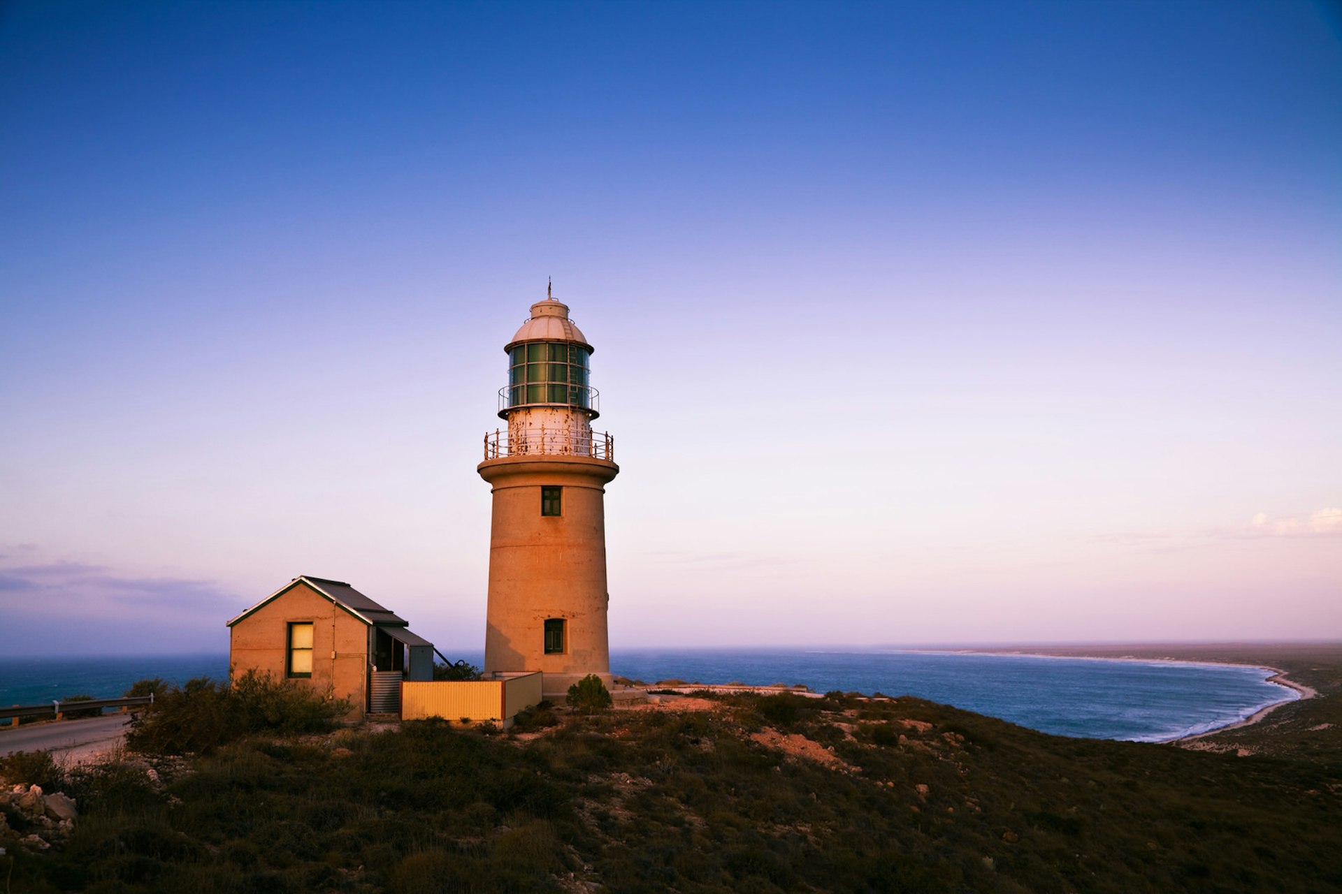 Ningaloo Lighthouse pictured during twilight; it sits atop a brush-covered hill, with the beach-lined coast curving away into the distance below