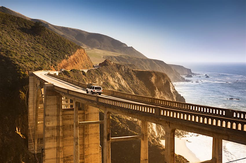 The sun highlights interesting architectural arches on Bixby Bridge as a car crosses with the ocean in the background near Monterey, California