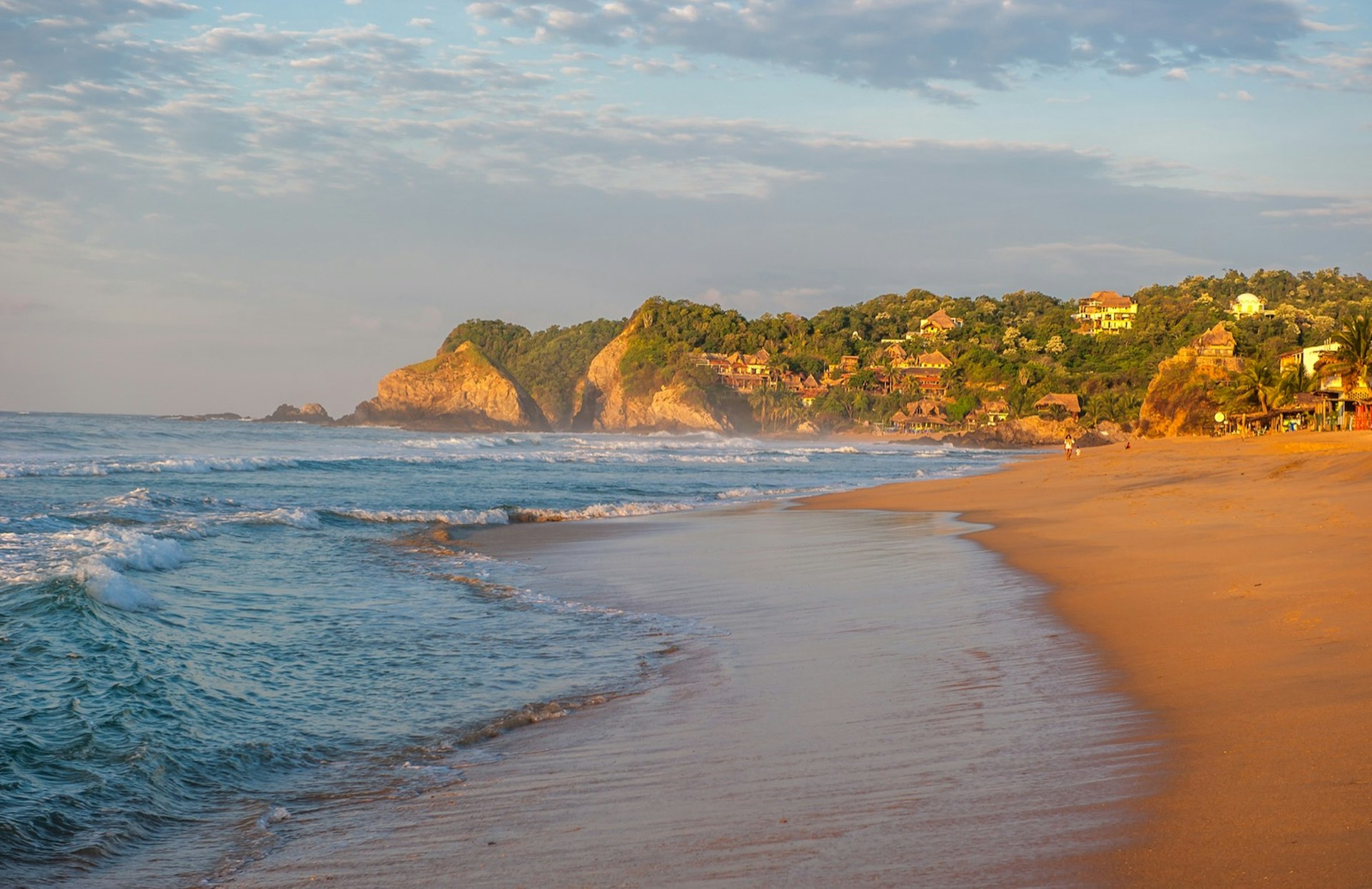 An empty white sand beach stretches to low green hills in the background with the ocean lapping against the shore