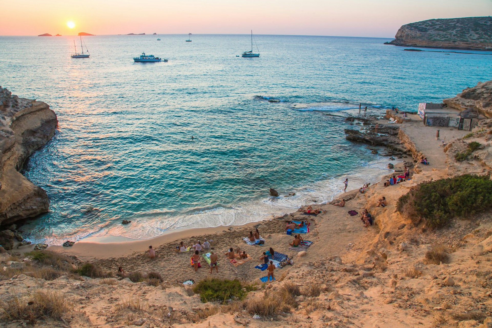 People line the sand at Cala Compte beach, Ibiza. A number of people sit on coloured towels while a couple swim in the sea. The sun sets in the background.
