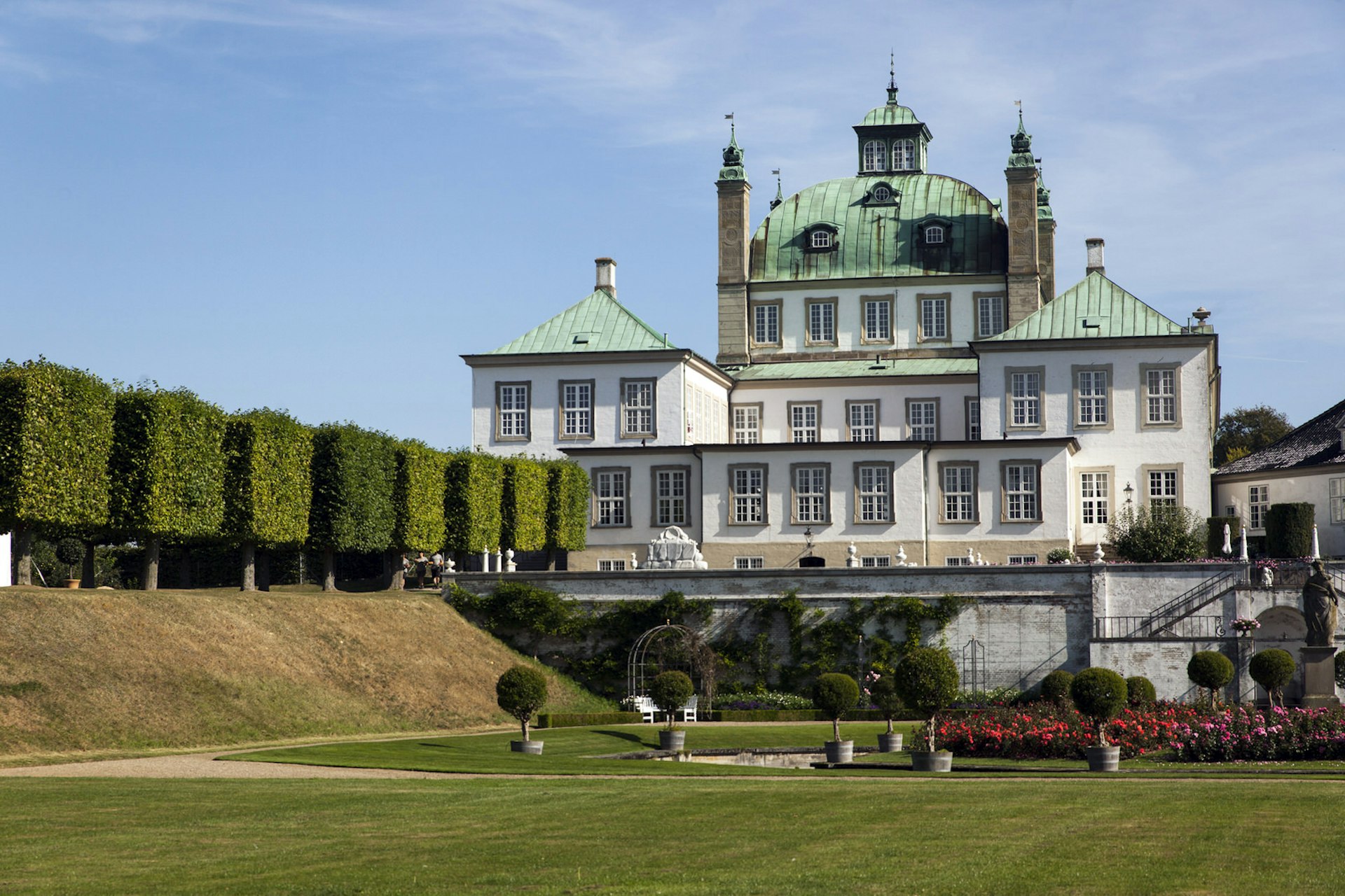 A green manicured lawn and row of large, square-shaped trees lead up to the rather grand Fredensborg Palace, with its white walls and green copper roof