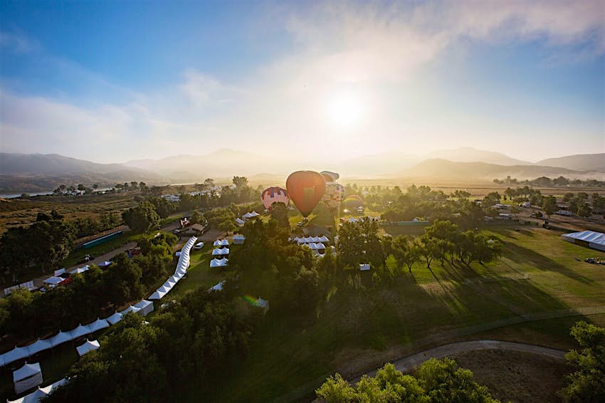 The sun rises over a verdant green valley while a red hot air balloon floats above in Temecula Valley at one of California's best summer food and drink festivals